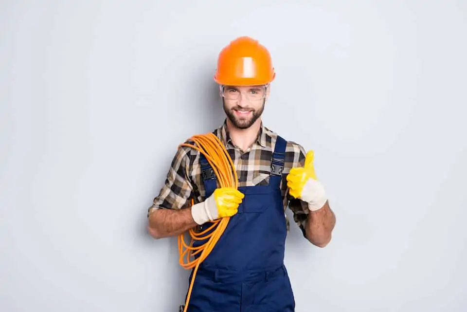 Portrait of handsome joyful electrician in hardhat, overall, shirt with bristle, holding rolled wires on shoulder, showing thumb up recommend approve sign over grey background; how to become an electrician