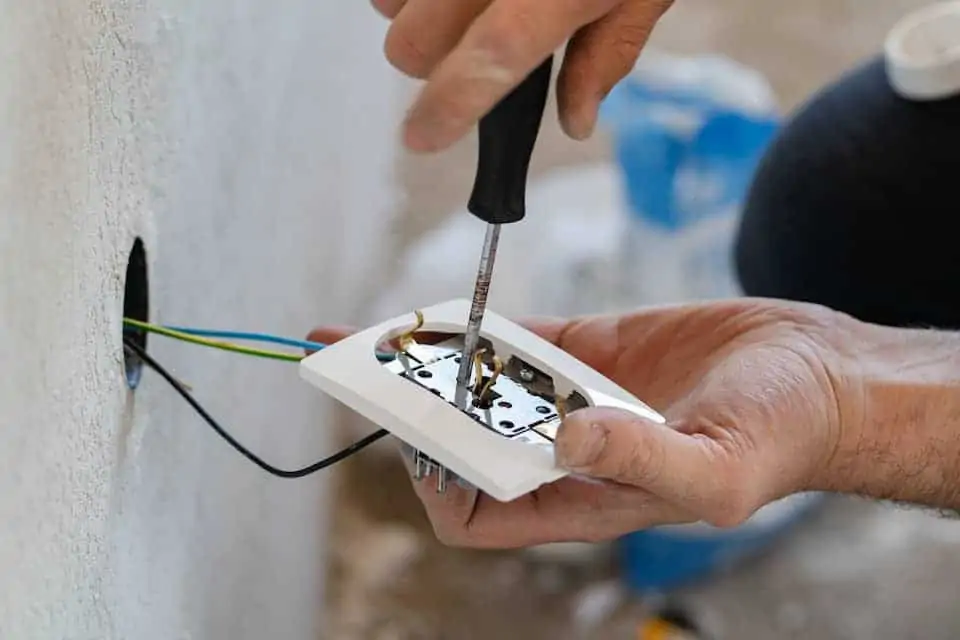 Close up on hands of caucasian man electrician holding screwdriver working on the plug electric on residential electric system installing white AC power socket on gray wall at home repair close up: outdoor diy outlet not working