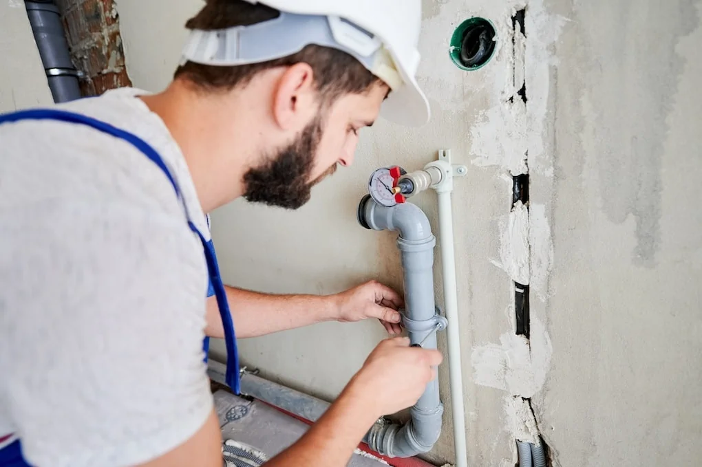 Close up of bearded young man in safety helmet screwing clamp on heating pipe with screwdriver; minnesota plumbing code