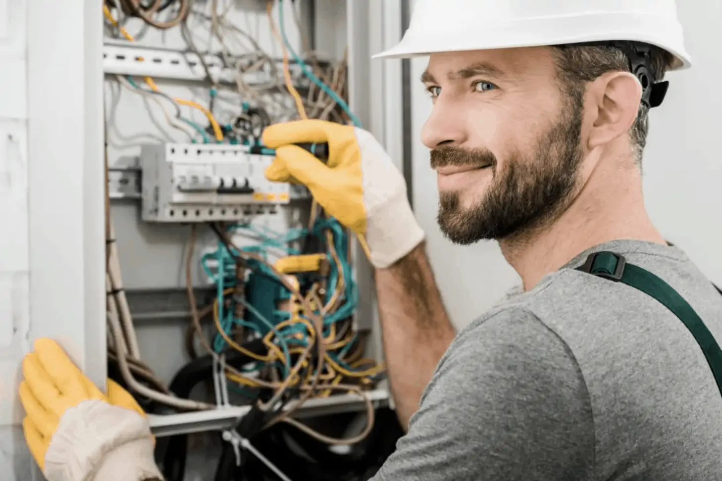 An electrician in a white construction hat and gloves, working on an electrical panel & smiling.