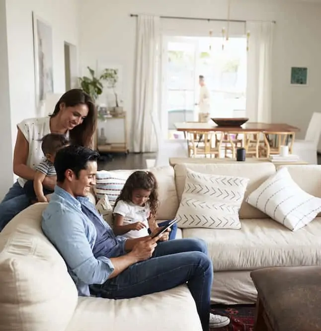 A family of four sitting on a light-colored couch and looking at a tablet in one parent's hand.