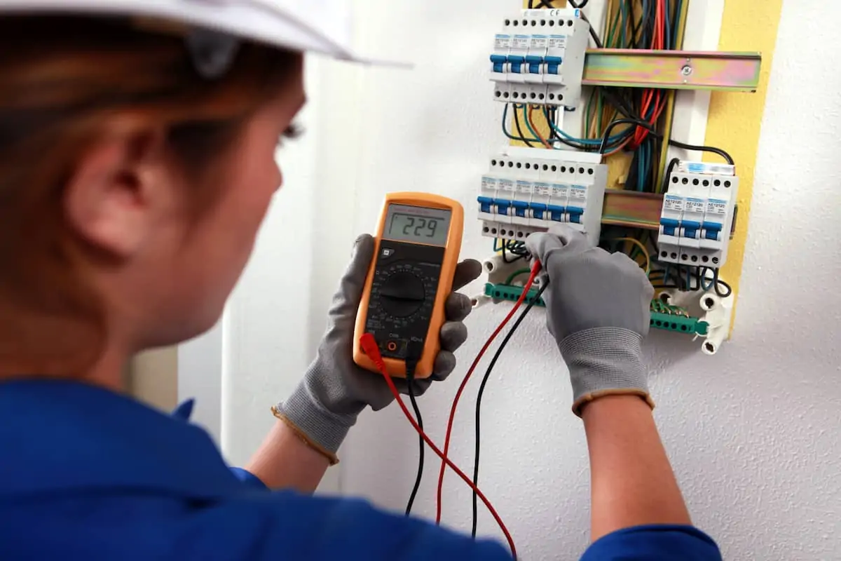 An electrician with a construction hat and gloves on tests voltage with a tool.