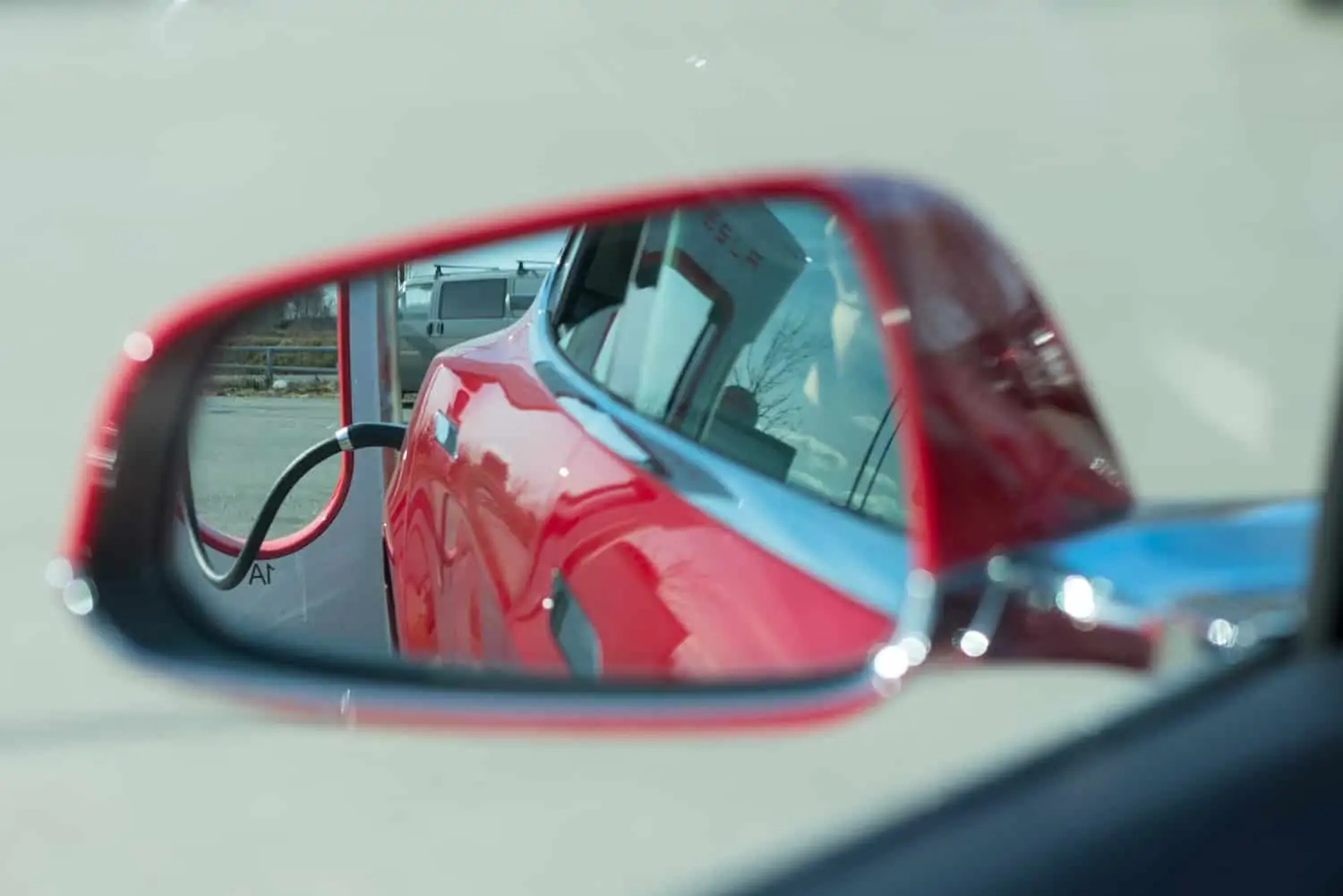 In the side rearview mirror of a red electric vehicle, there is a view of an attached EV charger.