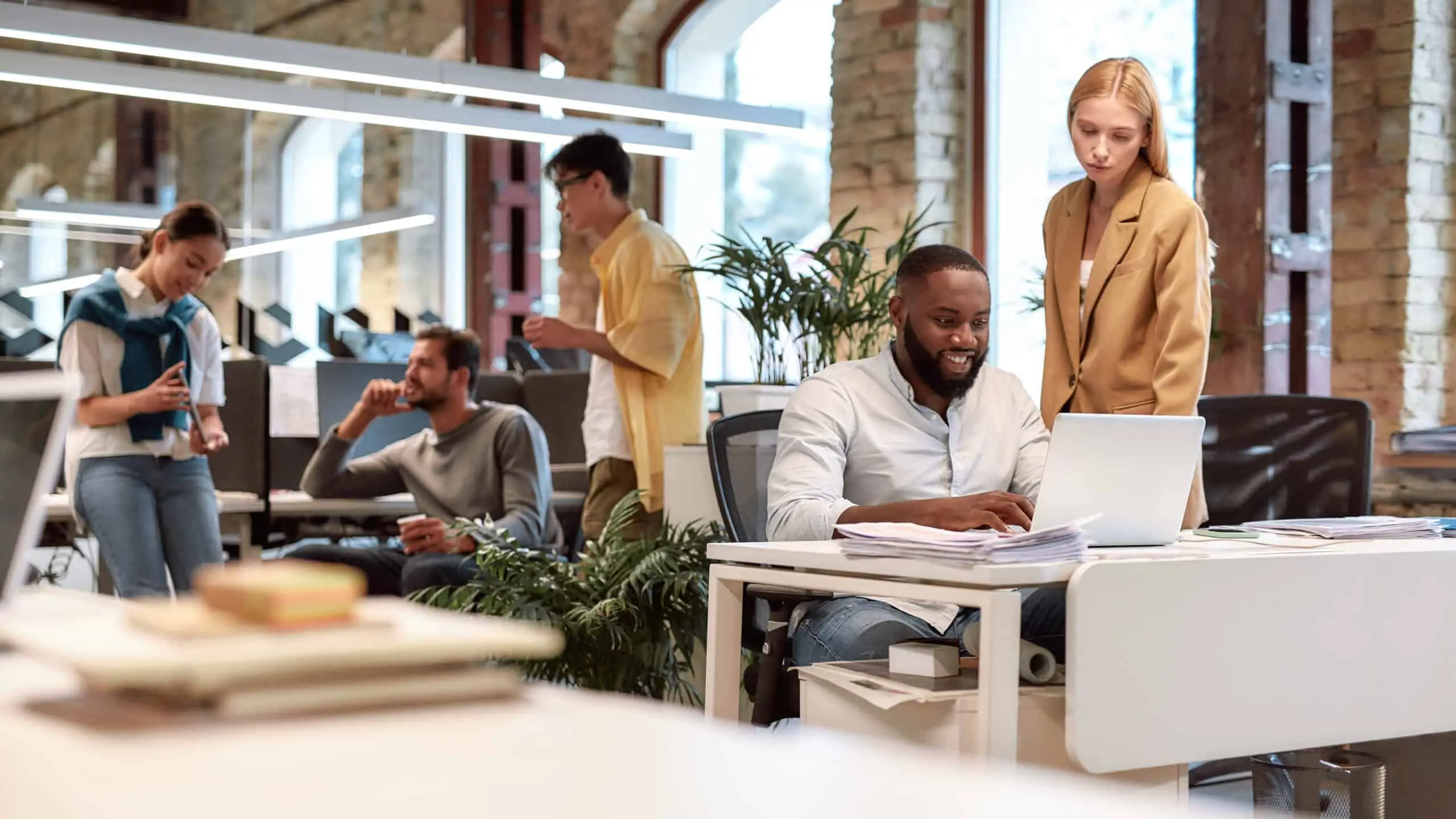 Coworkers in a casual office space—three of them look at a tablet & two of them look at a laptop.