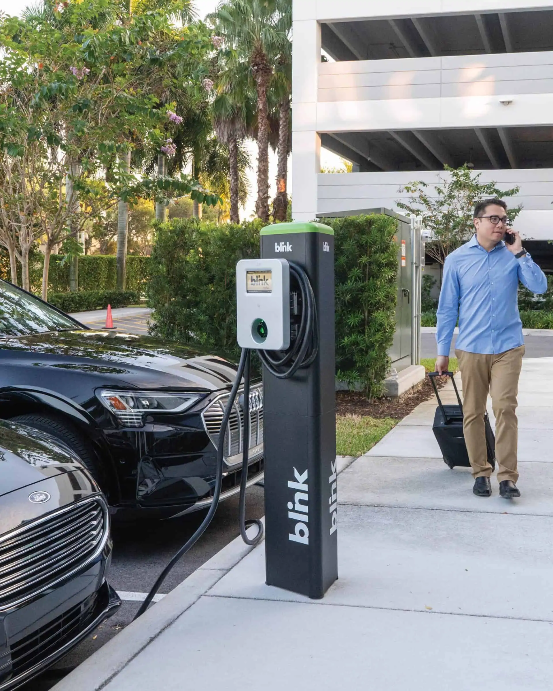 A view of a Blink charging system in a parking lot as a person with a rolling suitcase walks by.