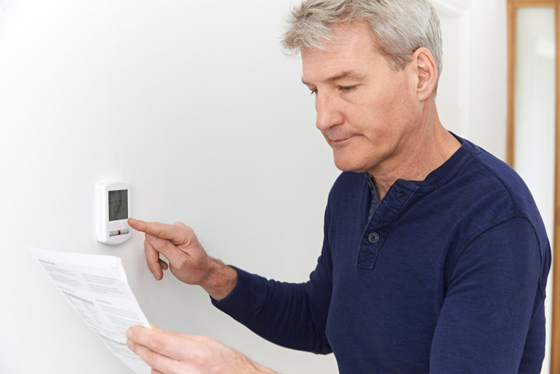 A person in a long-sleeved blue shirt reads instructions & pushes a button on a thermostat.