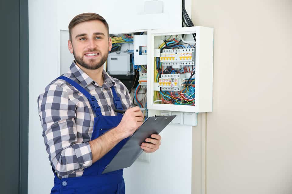 Male electrician with clipboard near fuse board indoors, space for text; how to become an electrician; amps vs watts vs volts