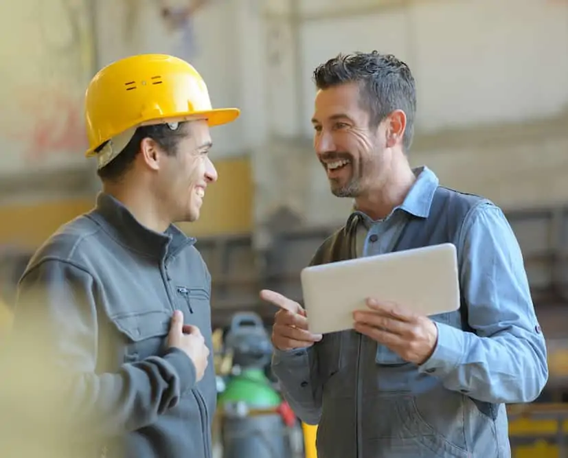 Two coworkers stand & smile at one another as they converse—one of them holds a tablet in hand.