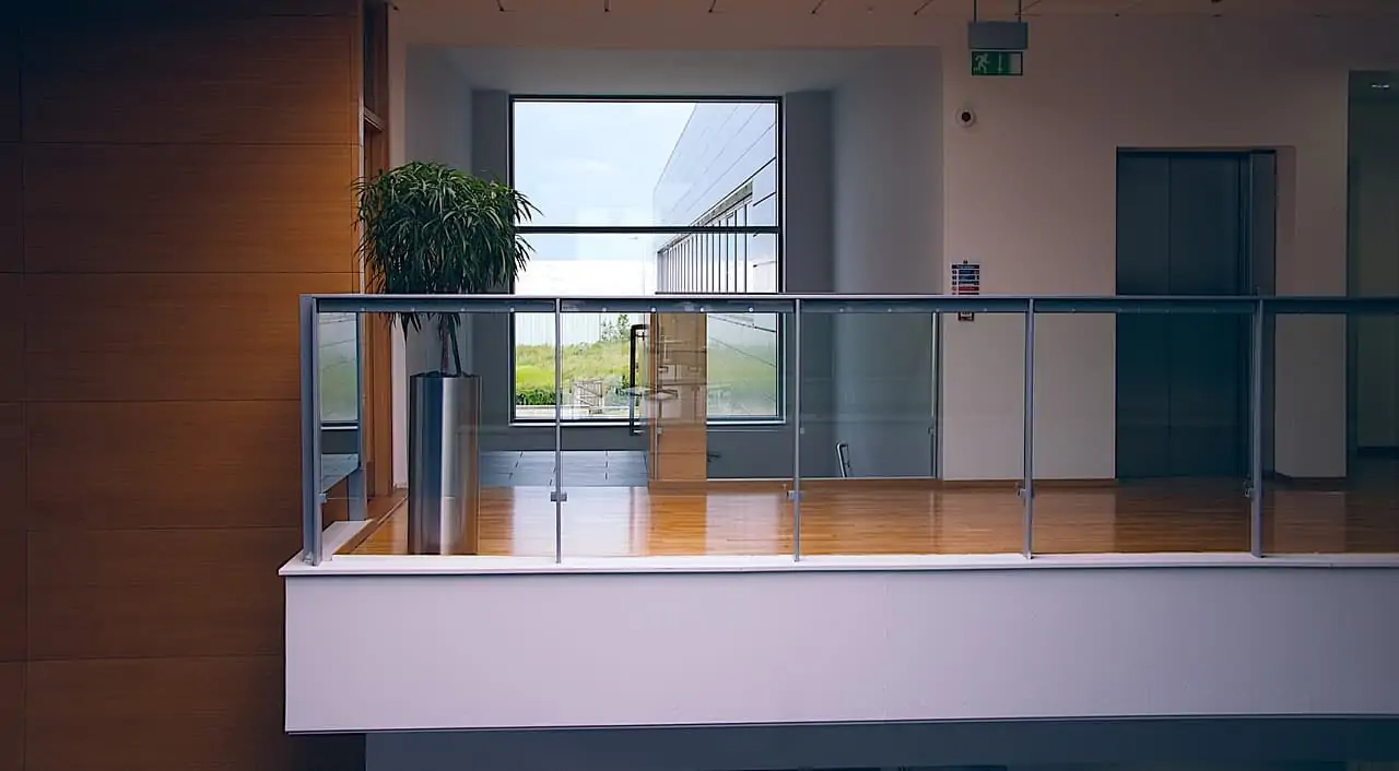 A modern building floor with glass railings, light brown wooden floors, a plant, and an elevator.